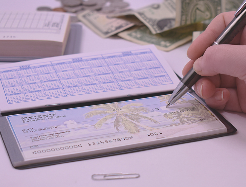 A lifestyle shot of a person filling out a tropical-themed check amidst a desk setting with cash, coins, and a calendar insert.